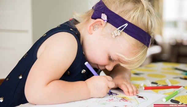 A toddler wears hearing aids attached to a headband while drawing.